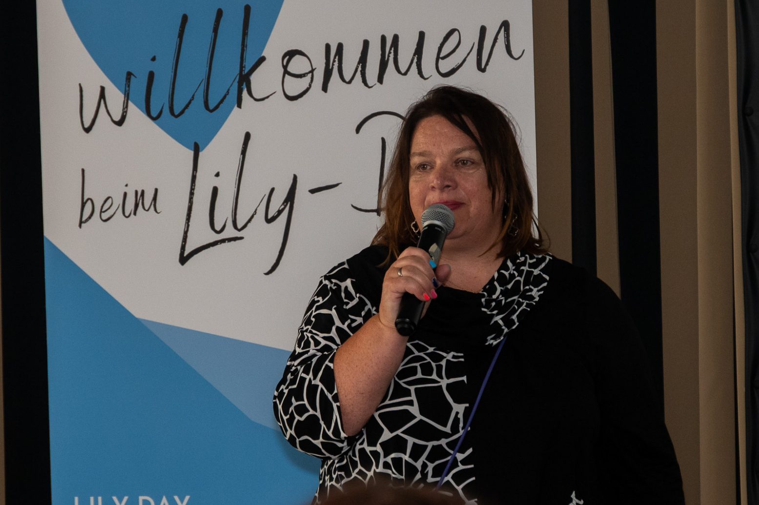 Lily-day Frau spricht auf einer Veranstaltung, mit einem Mikrofon in der Hand, im Hintergrund ein Banner.